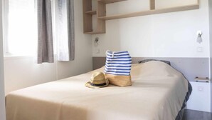 A bed with a striped pillow and a straw hat, a wooden shelf above it, and a window with curtains. - Cap - Confort 2 TV - 4/5p (15) (Plouézec)