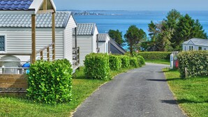 A row of white mobile homes with grey roofs, a paved road, and a view of the sea. - Cap - Confort 2 TV - 4/5p (15) (Plouézec)