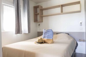 A bed with a striped pillow, a straw hat, and a striped bag. There are wooden shelves above the bed and a window with curtains. - Cap - Confort 2 TV - 4/5p (21) (Plouézec)