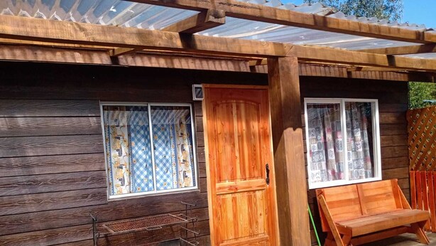 Una cabaña de madera con puerta corredera, ventana con cortinas, parrilla metálica y banco de madera.