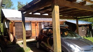 Un coche estacionado bajo una estructura de madera con techo metálico, junto a un edificio de madera con ventana y puerta.