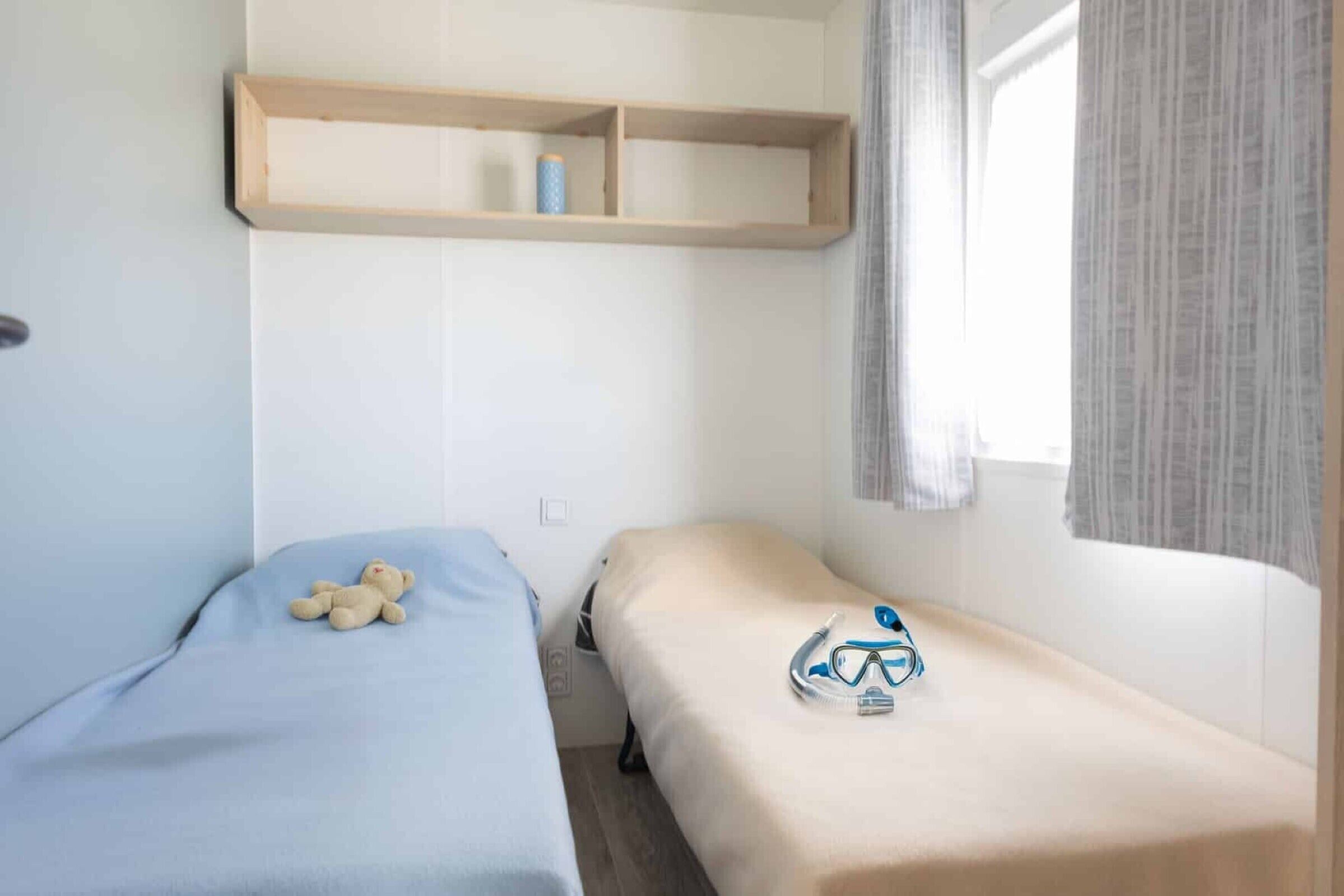 A bedroom with two beds, a shelf above, and a window with curtains.