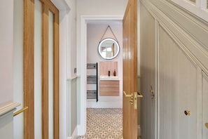 A hallway leading to a bathroom with a patterned floor, a round mirror, and a wooden door with a brass handle. - Stylish 4 bed home in Old Trafford next to MUFC (Stretford)
