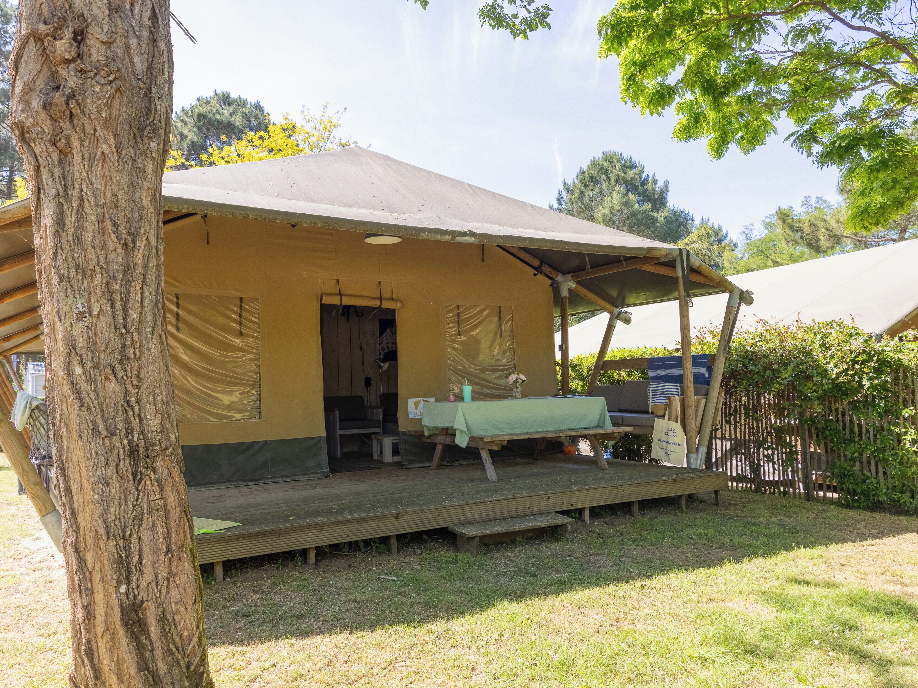 A rustic outdoor accommodation with a covered seating area, a table, and a bench.