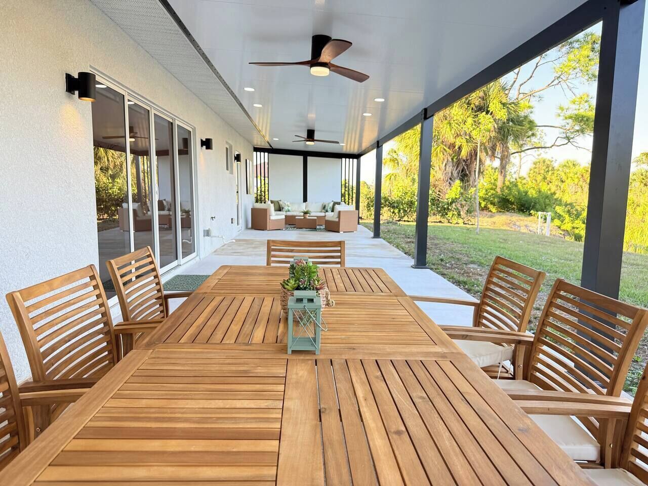 Eine überdachte Terrasse mit Holztisch und -stühlen, großen Fenstern und Deckenventilatoren.