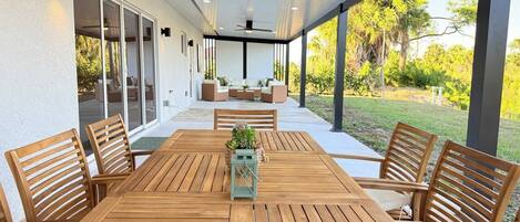 Eine überdachte Terrasse mit Holztisch und -stühlen, großen Fenstern und Deckenventilatoren.