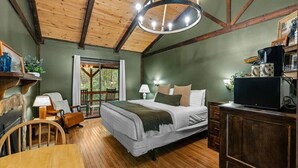 Un dormitorio con techo de madera, una cama con ropa blanca, un escritorio de madera con una lámpara y un televisor sobre un mueble.