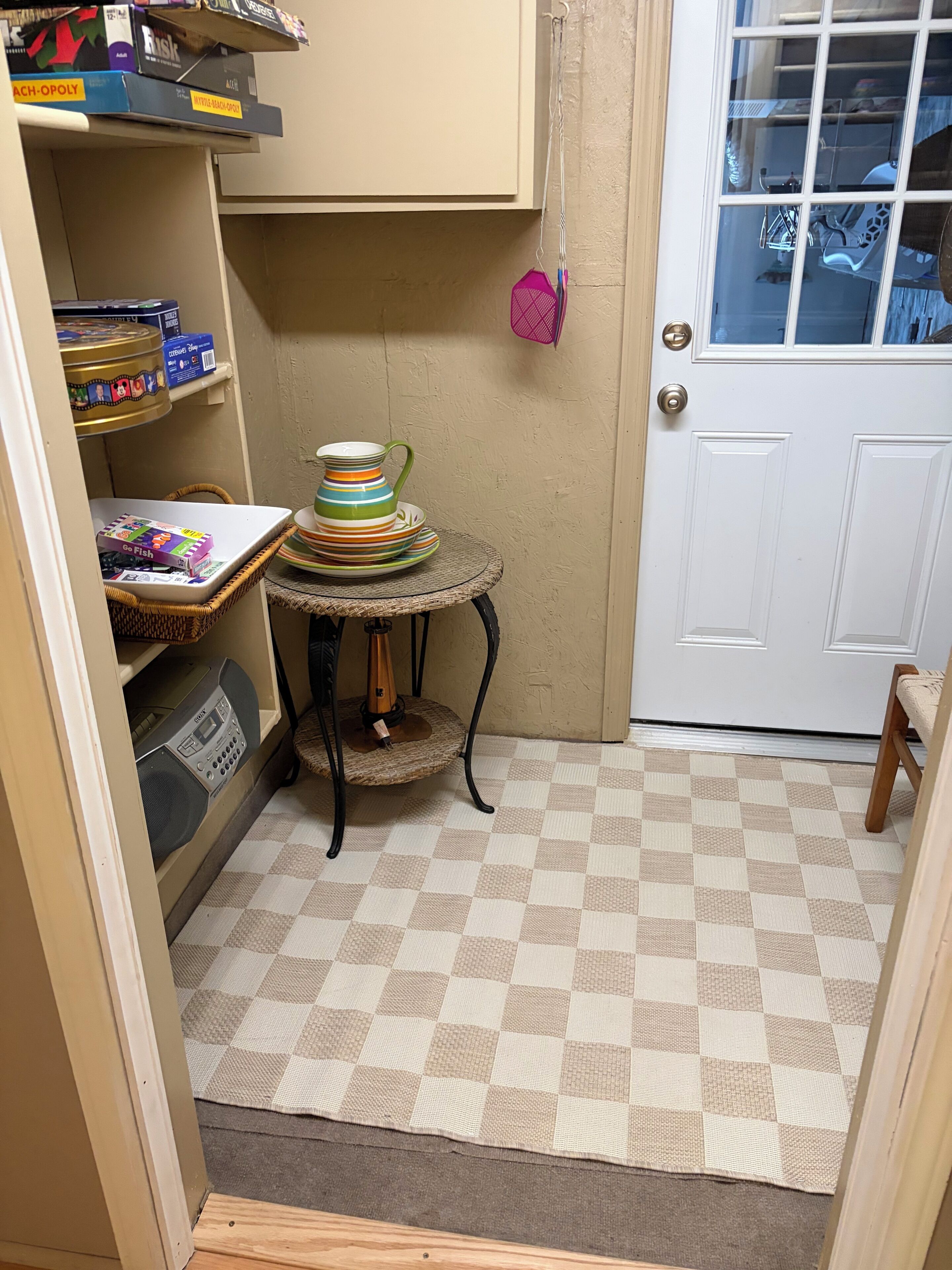 A small room with a checkered floor, a white door, a wicker table with a radio, and shelves with various items.
