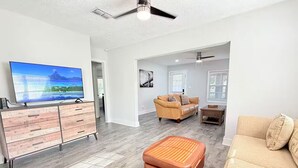 Ein geräumiges Wohnzimmer mit einem großen Flachbildfernseher, einer Holzkommode, einem braunen Hocker, einem beigen Sofa und einem Deckenventilator.