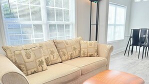 Ein beiges Ecksofa mit Kissen im Palmenmuster, ein brauner Ledersitzhocker und ein Essbereich mit schwarzen Stühlen und einem Glastisch.