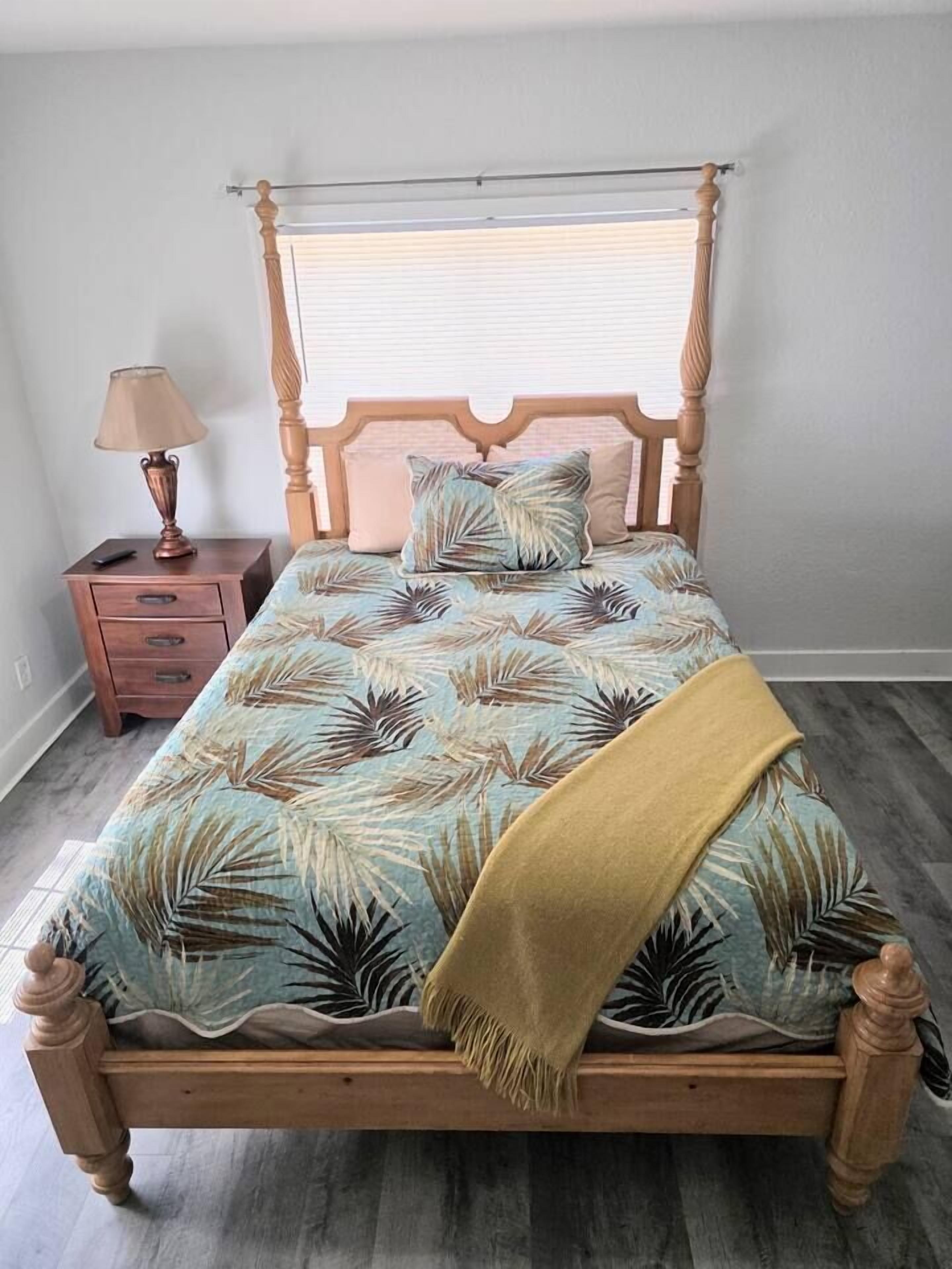 A bed with a wooden frame, a patterned bedspread, a bedside table with a lamp, and a window with blinds.