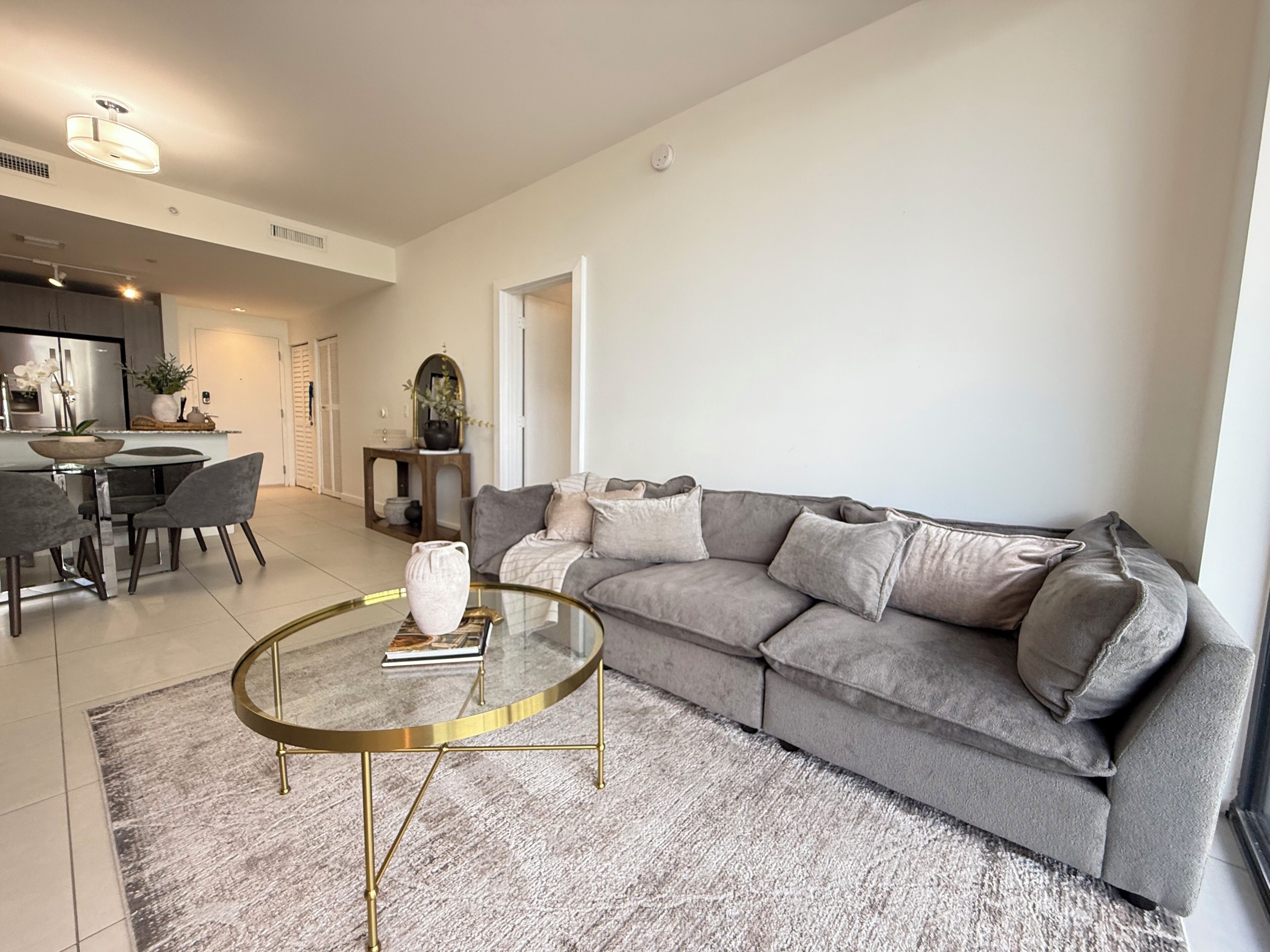 Ein modernes Wohnzimmer mit einem grauen Sofa, einem Glastisch und einem Essbereich mit weißem Tisch und Stühlen.
