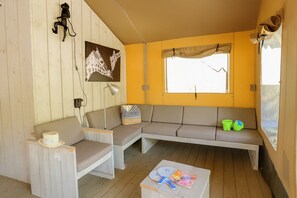 Ein gemütliches Zimmer mit Holzboden, einer Couch, einem Sessel, einem kleinen Tisch mit Hut und Zeitschrift sowie einem Fenster mit Vorhang.