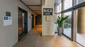 A modern hallway with an elevator, a sign for 'NEX ONE Residencial', and a potted plant. - Edifício Nex One by Wecare Hosting (São Paulo)