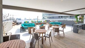 A rooftop terrace with wooden flooring, a teal sofa, and a bar area. - Edifício Nex One by Wecare Hosting (São Paulo)