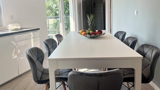Een moderne eethoek met een witte tafel en zes beklede grijze stoelen. Er staat een fruitschaal op de tafel, en in de achtergrond is een koelkast zichtbaar.