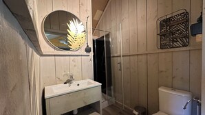 Un baño con pared de paneles de madera, un espejo redondo, un lavabo blanco, cabina de ducha y un inodoro.