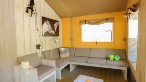A cozy room with a wooden floor, a sofa, a chair, a small table with a hat and a magazine, and a window with a curtain. - Palmyre - Eco-lodge 3 - 7p (10) (Les Mathes)