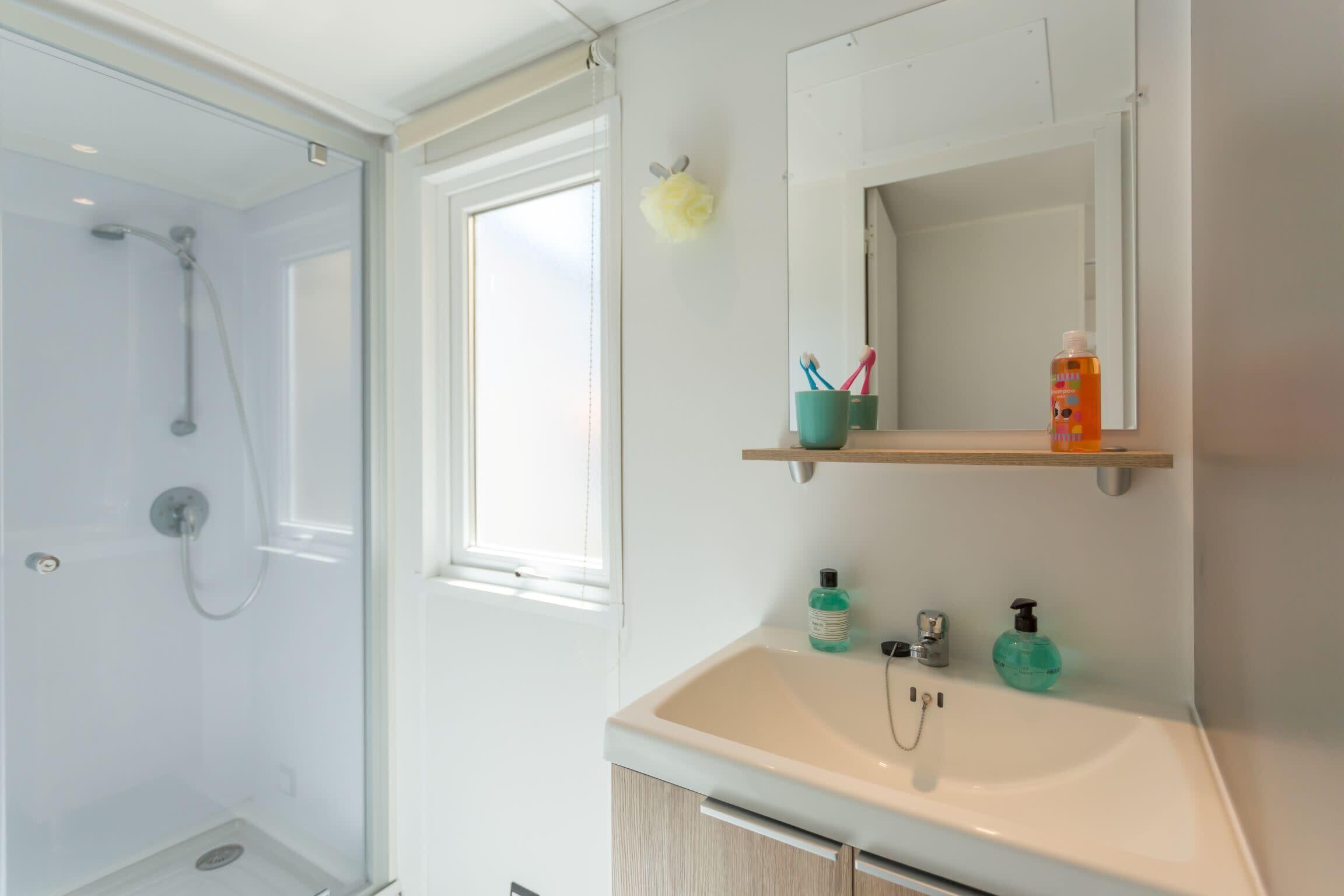 A bathroom with a glass-enclosed shower, a white sink with a mirror above it, and a window providing natural light.