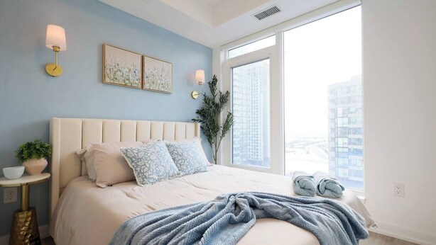 A bedroom with a large bed, a window with a city view, a bedside table with a lamp, and a potted plant. - The Calm House | 5 mins Walk to Square One Mall (Mississauga)