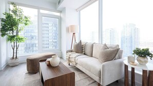 A modern living room with a sofa, a coffee table, and a floor lamp. There is a large window with a view of high-rise buildings. - The Calm House | 5 mins Walk to Square One Mall (Mississauga)