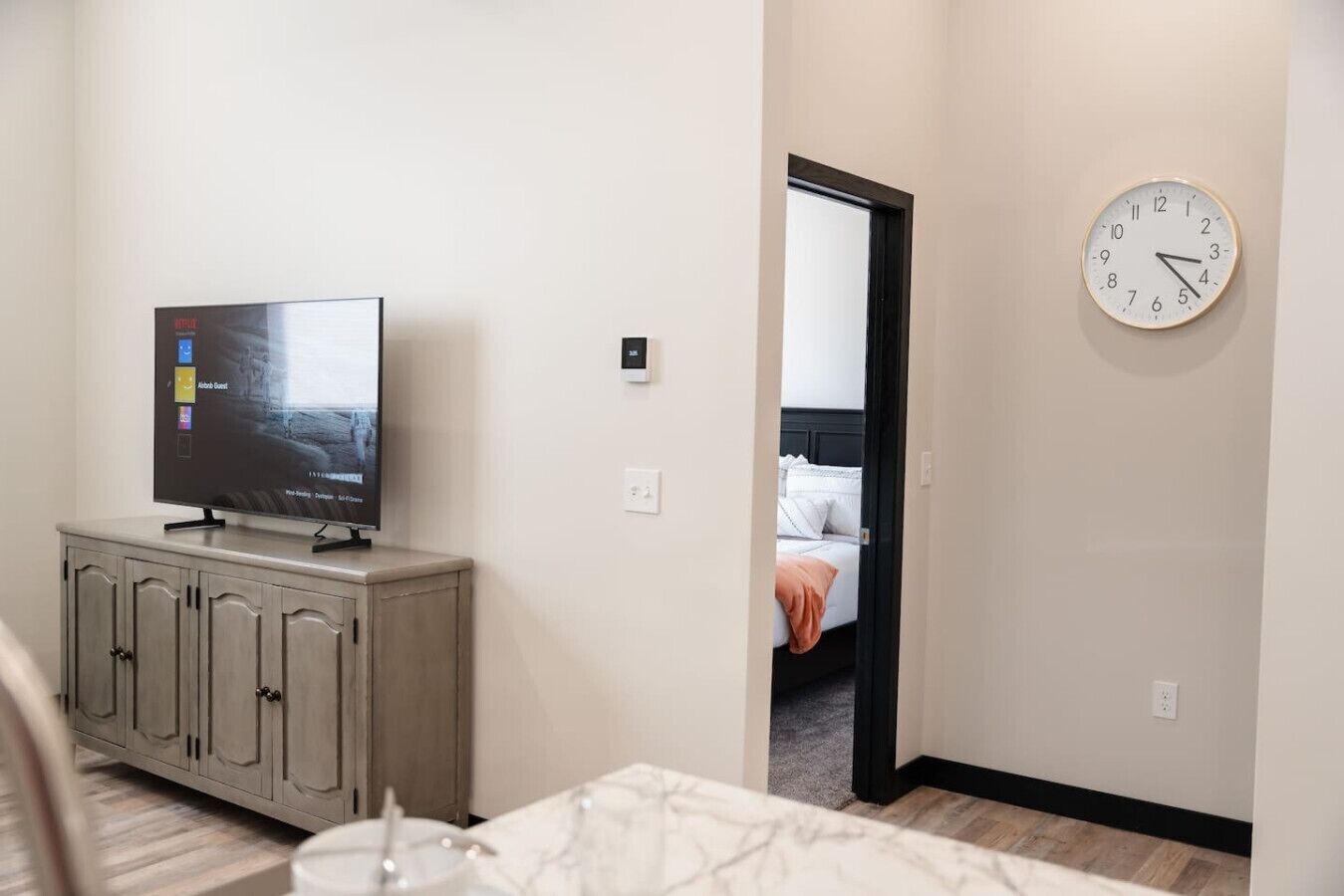 A hotel room with a television on a cabinet, a clock on the wall, and a bed with a white headboard.