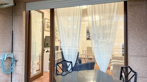 Una habitación con una puerta corrediza de vidrio que da a un balcón, una mesa de comedor con sillas y vistas del interior a través de cortinas traslúcidas.