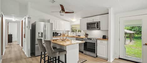 A modern kitchen with white cabinetry, a stainless steel refrigerator, a microwave, an oven, and a bar area with two stools.