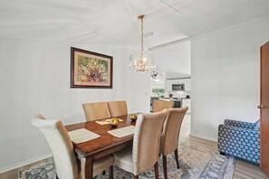 A dining area with a wooden table, beige chairs, a chandelier, and a kitchen in the background.