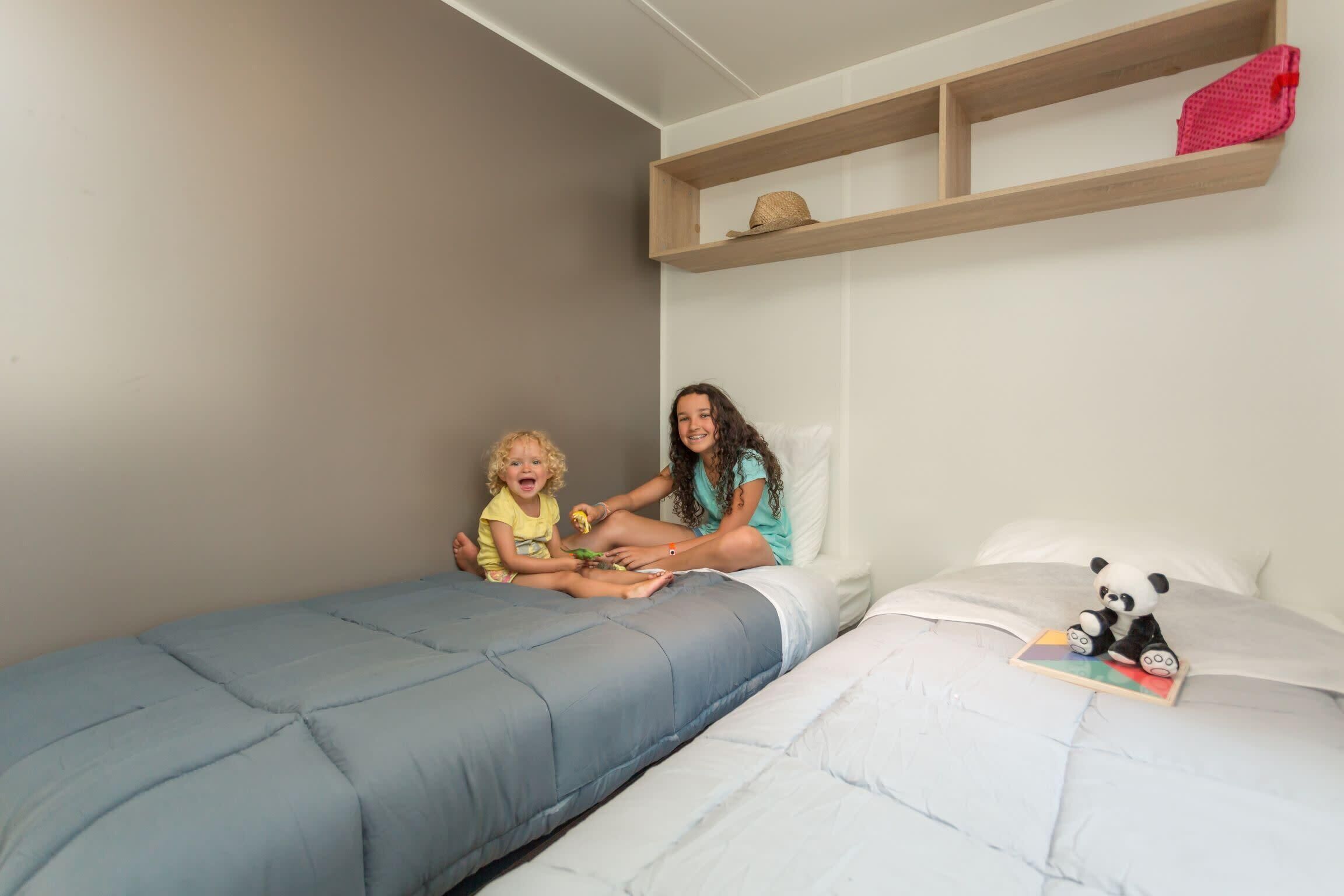 Un dormitorio con dos camas, una con colcha azul y la otra con colcha blanca. Hay un estante sobre las camas con una cesta y un objeto rojo.