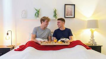 Duas pessoas sentadas em uma cama com itens de café da manhã, uma luminária, e uma parede com um quadro e plantas.