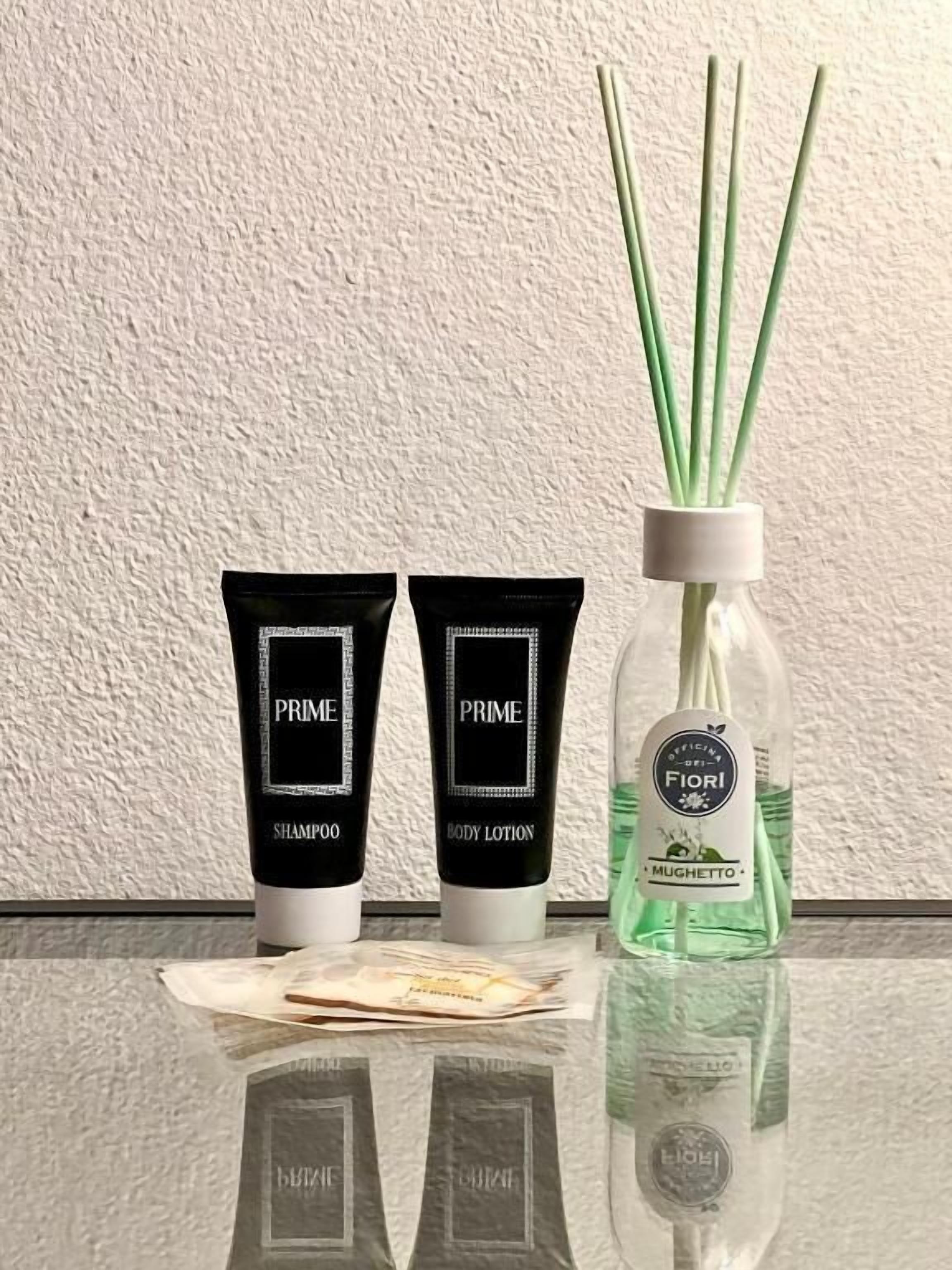 Two black tubes labeled 'PRIME SHAMPOO' and 'PRIME BODY LOTION' and a clear bottle with green reeds labeled 'Fiori MUGHETTO'.