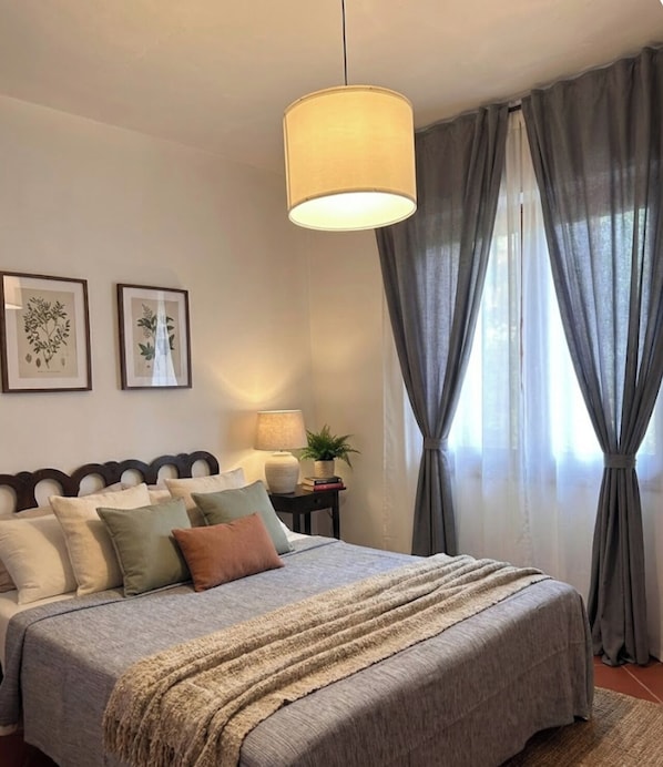 A bedroom with a bed, a bedside table with a lamp, a plant, and two framed botanical prints on the wall. - B&B RIO GRANDE (Massarosa)