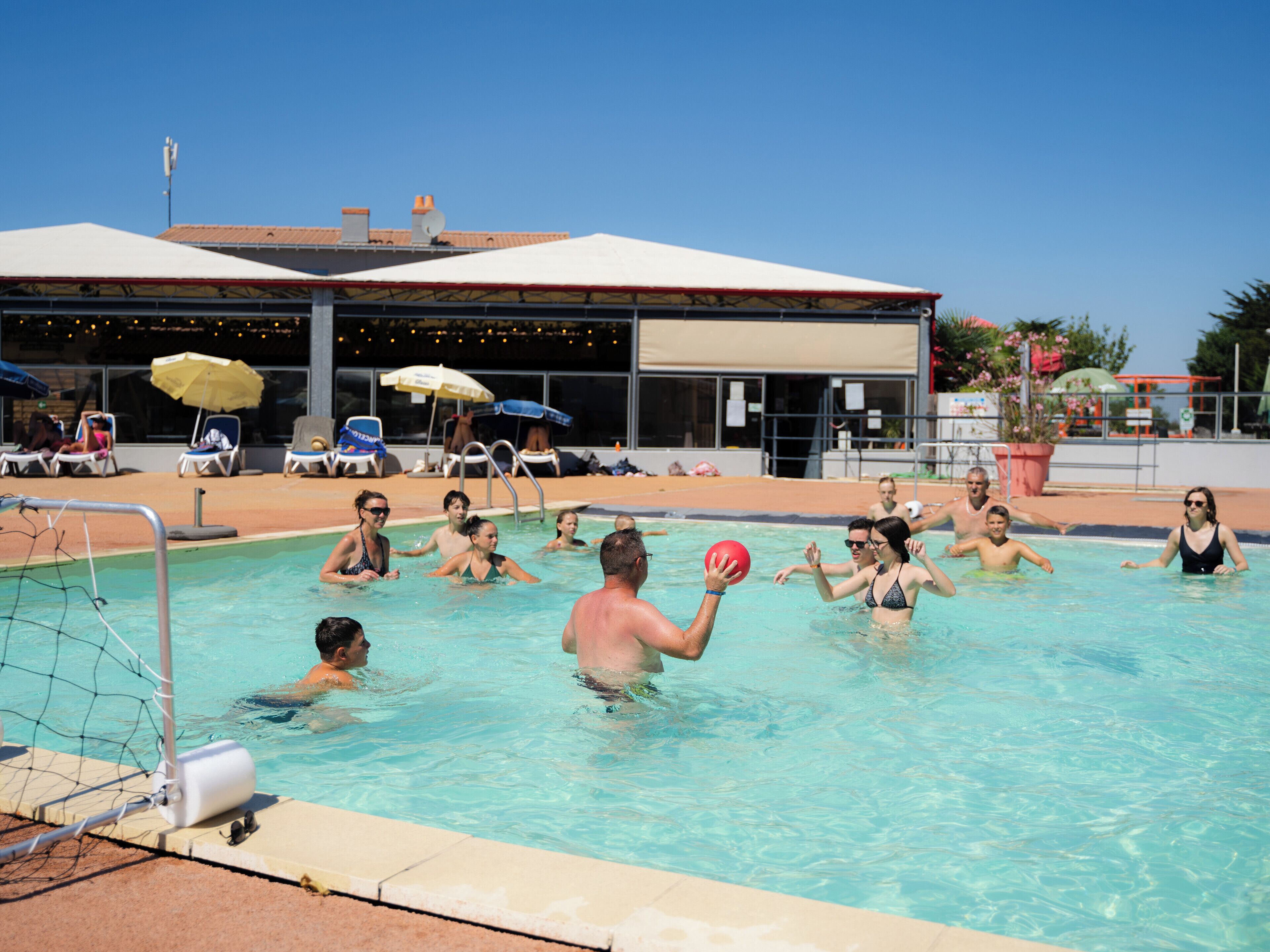 Ein Schwimmbecken mit spielenden Personen, Liegestühlen und Sonnenschirmen.