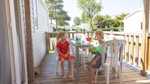 Twee kinderen zitten aan een tafel op een houten terras met witte stoelen.