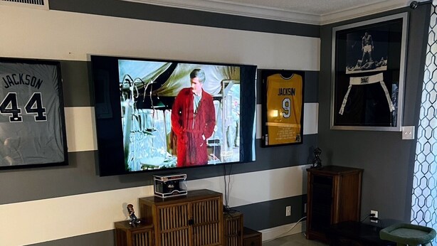 Un salón con un televisor de pantalla plana montado en la pared, un sillón blanco, un mueble de entretenimiento de madera y un rascador para gatos verde.