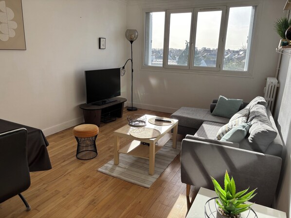 A modern living room with a flat-screen TV, a sofa, a coffee table, and a view of the city through large windows. - Bright and quiet apartment close to the center of Rennes and the train station. (Rennes)