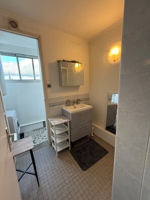A compact bathroom with a white sink, mirror, and a small vanity. There is a window with a view of the sky and clouds, and a door leading to another room. - Bright and quiet apartment close to the center of Rennes and the train station. (Rennes)