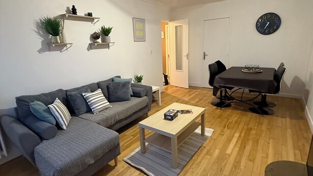 Ein Wohnzimmer mit grauem Sofa, einem Couchtisch, einem Esstisch mit Stühlen und einer Wanduhr.