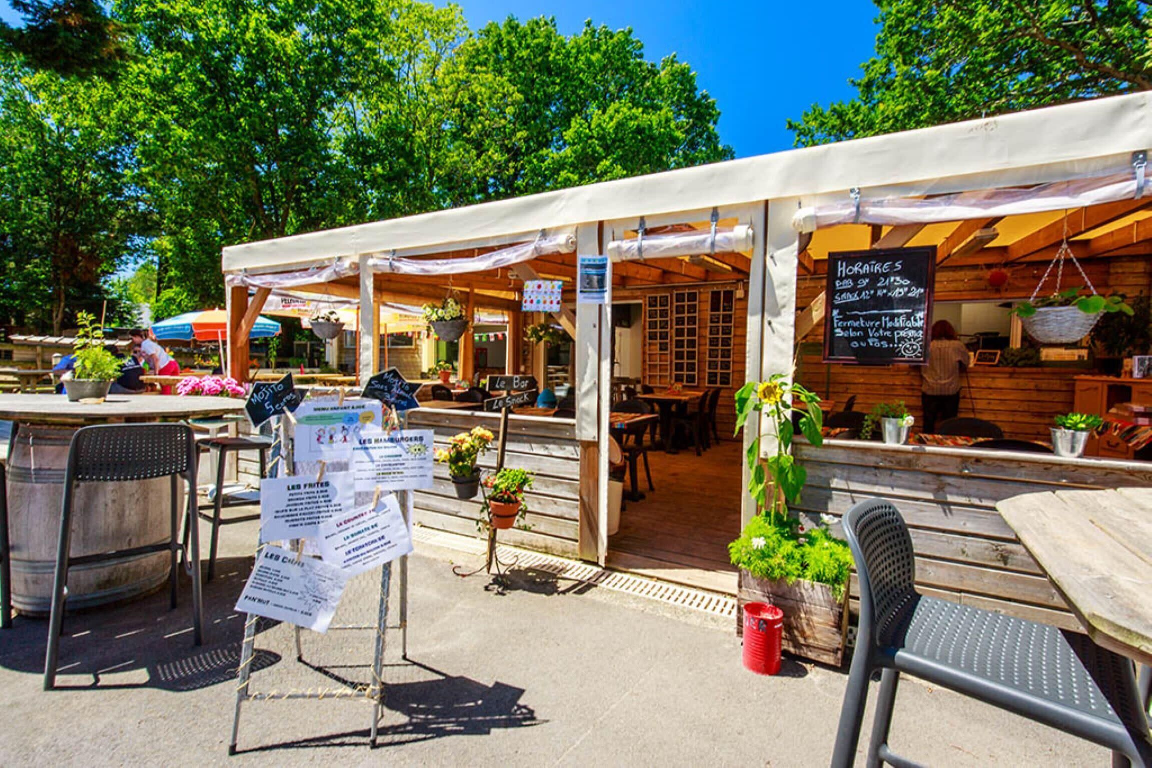 Un café al aire libre con mesas, sillas, un tablero de menú y plantas en macetas.
