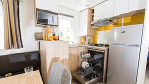 Een kleine keuken met een magnetron, kookplaat en vaatwasser. Er staat een eettafel met een stoel en een glas erop.