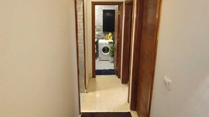 Un couloir étroit, recouvert d’un tapis noir, mène à une pièce où se trouvent une machine à laver et une plante.