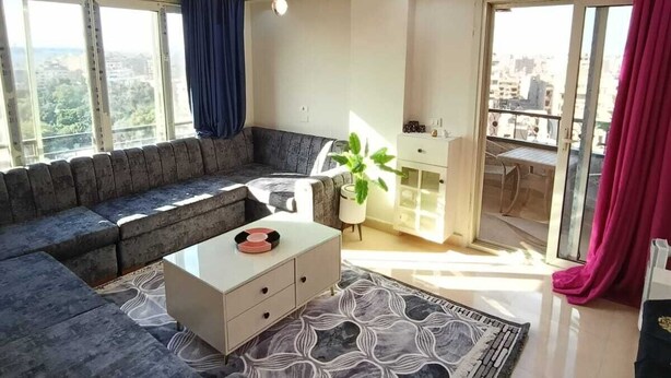 Un salon moderne avec un canapé d’angle gris, une table basse blanche et un tapis à motifs. Il y a un balcon avec vue sur la ville.