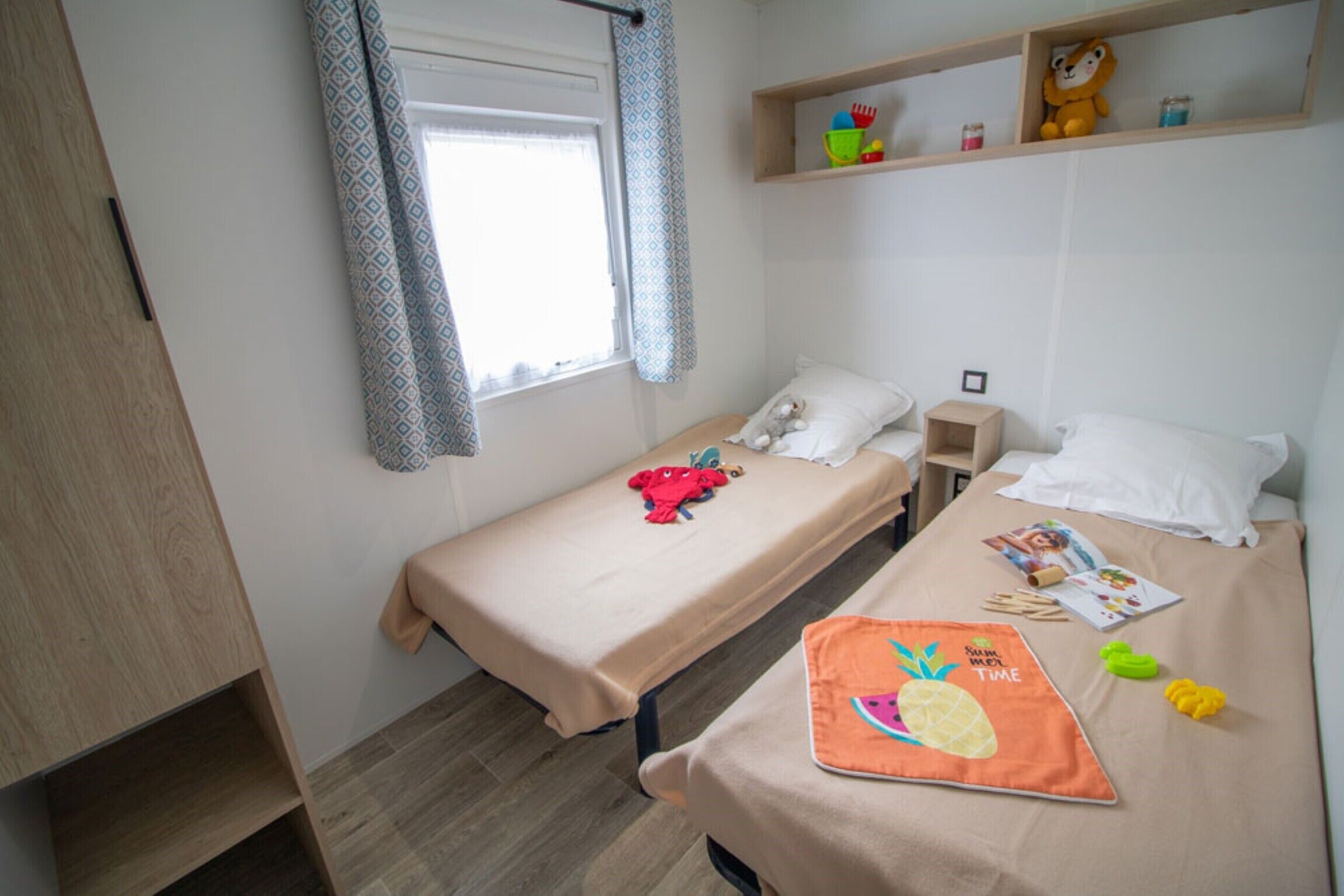 Une petite pièce bien éclairée, avec deux lits simples, une armoire en bois, une fenêtre ornée de rideaux à motifs et une étagère remplie de jouets.