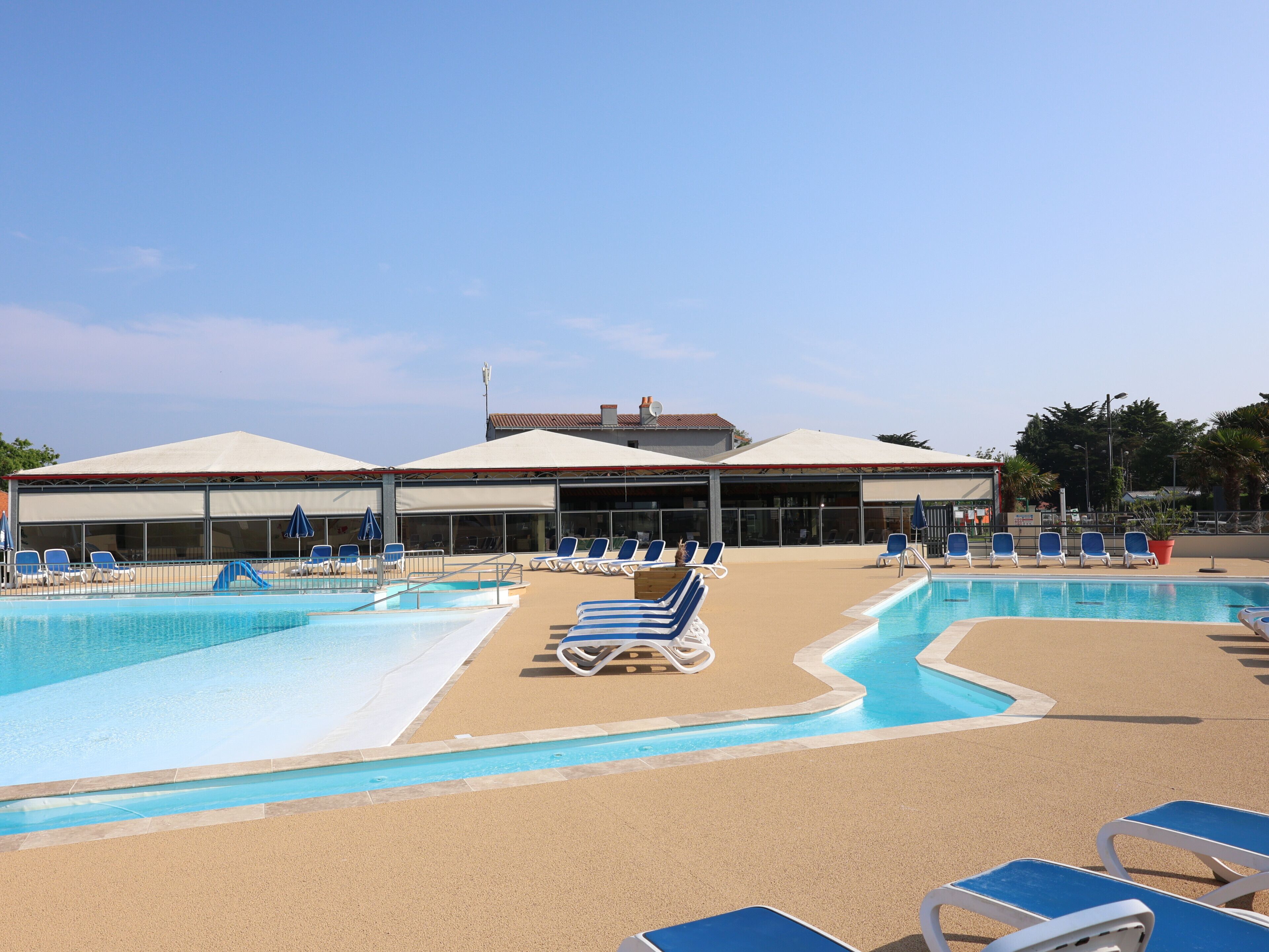 Un espace piscine doté de nombreuses chaises longues et de parasols, un bâtiment avec de grandes fenêtres et un toit abrité, et un ciel dégagé.