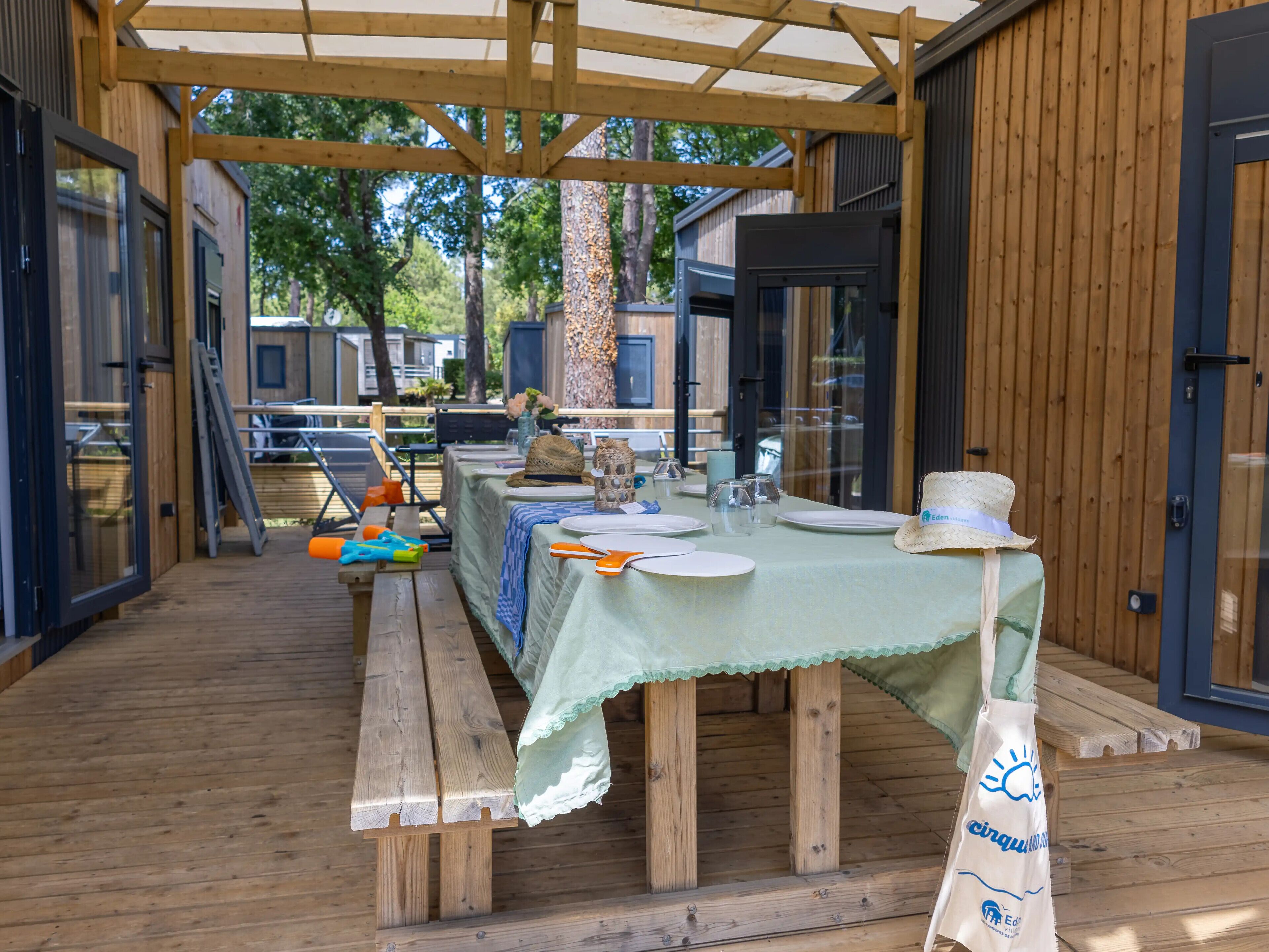 Een buitenterras met een houten dek, een overdekte tafel met tafelkleed en een picknickbank.