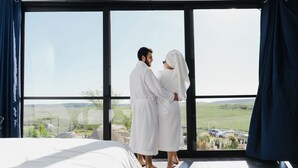 Un couple, vêtu de longues robes blanches, se tient près d’une grande fenêtre donnant sur un paysage magnifique.