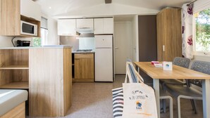 Una cocina compacta con armarios de madera, microondas, estufa y refrigerador. Hay una mesa de comedor con sillas y un banco. Se ve una bolsa de tela con texto en el piso.