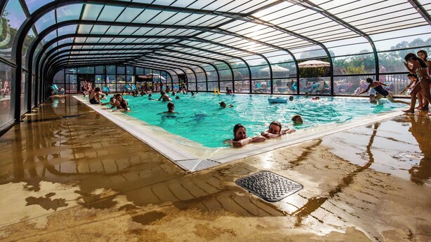 Una piscina techada con techo curvo, rodeada de gente nadando y tomando el sol.