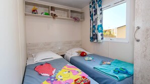 Ein Zimmer mit zwei Betten, einem fensterbelaubten Fenster, einem Regal mit Spielzeug und einer UNO-Kartenschachtel.